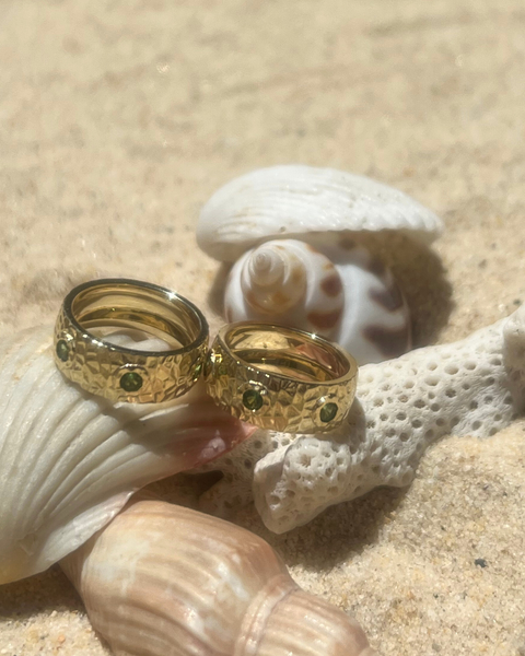 Two gold rings with green gemstones on a sandy surface with shells.